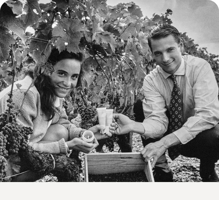 Two people smiling and kneeling between vineyard rows, holding soil samples beside a crate. Grape clusters and leafy vines surround them.