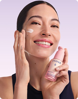 Smiling woman with glowing skin holding a pink skincare bottle against a lavender background