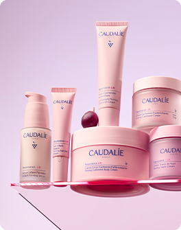Caudalie skincare products arranged on pink background, including bottles, tubes, and jars with pink and white packaging and a plum fruit.