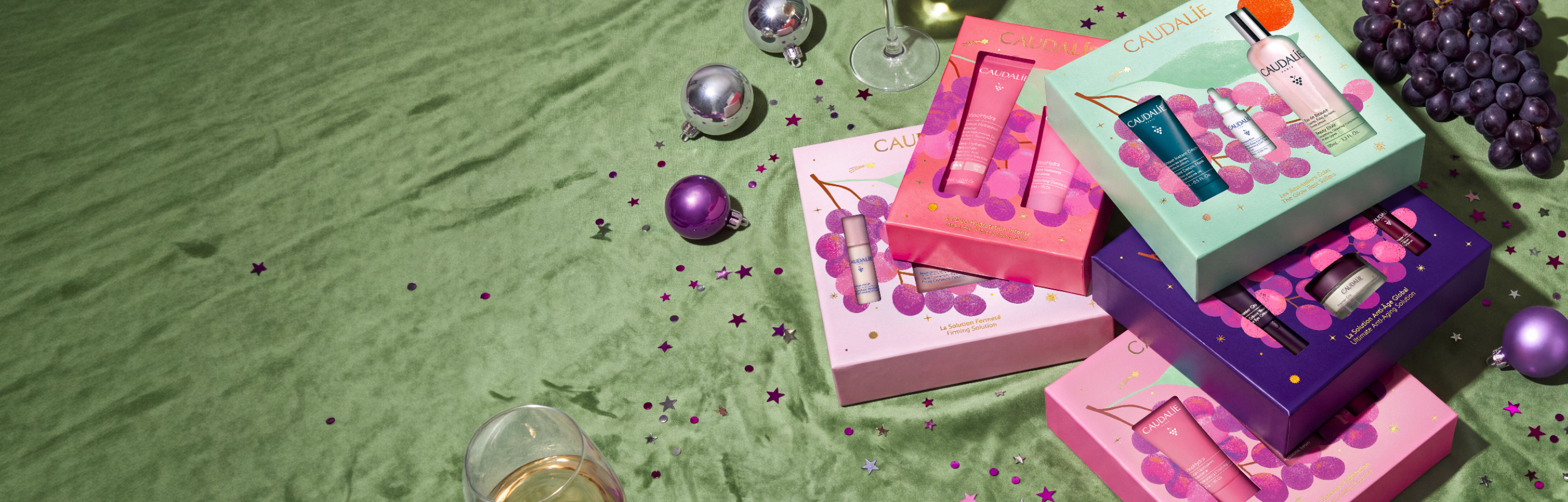 Caudalie beauty gift sets arranged on green textile with purple ornaments, grapes, and star confetti in festive holiday display.